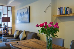Un salón con una mesa de madera, un jarrón de rosas rosadas, un sofá marrón y una estantería llena de libros.