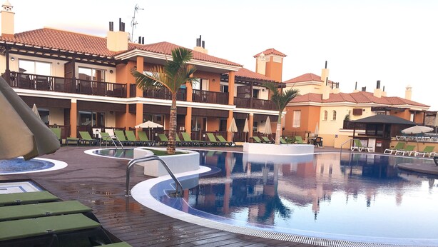 A pool area with lounge chairs and umbrellas, surrounded by buildings with balconies and a tiled roof. - Monte Carrera The Little Mermaid (Arguineguin)