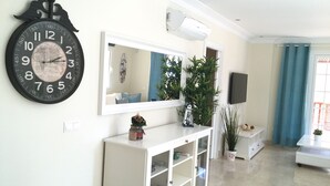 A living room with a large wall clock, a flat-screen TV, a white console table, and a potted plant. - Monte Carrera The Little Mermaid (Arguineguin)