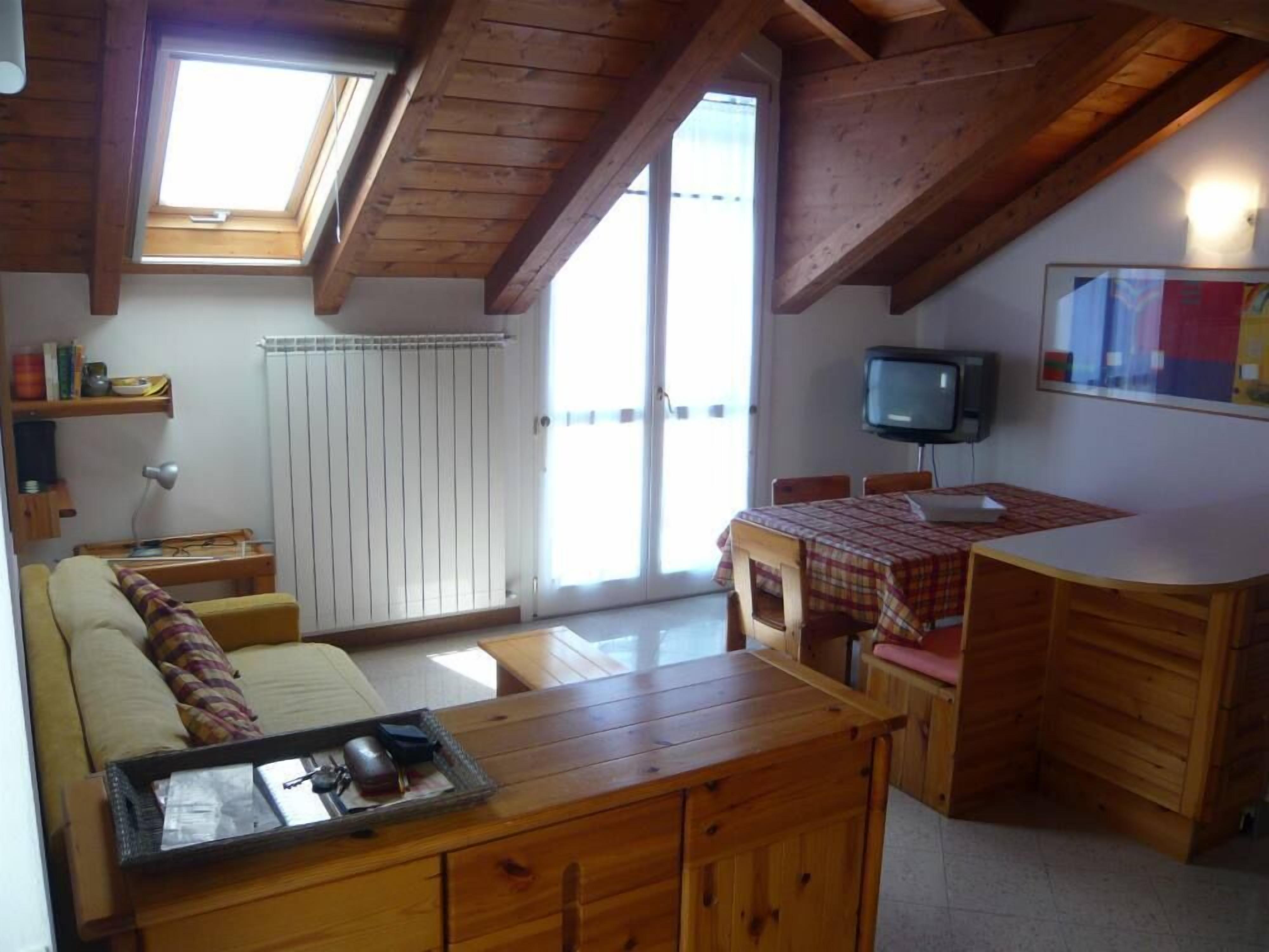 Een gezellig chaletinterieur met een houten eettafel, een bank, een televisie en een raam met jaloezieën.