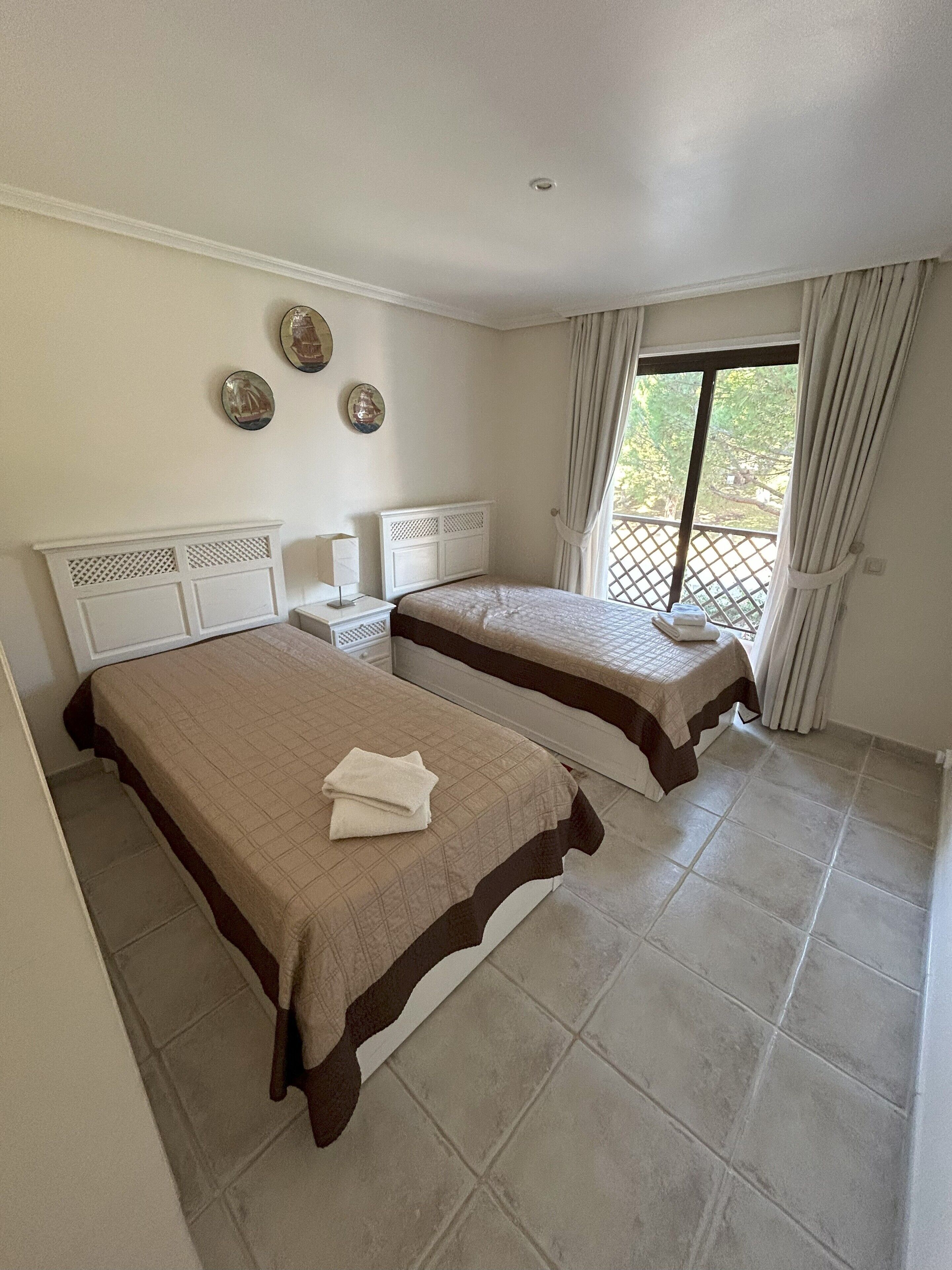 A hotel room with two beds, a nightstand, a lamp, and a window with curtains.