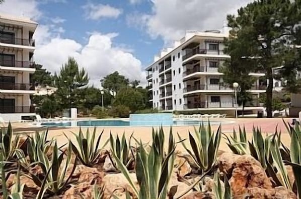 A pool area with a clear sky, surrounded by multi-story buildings with balconies, and some greenery in the foreground. - Luxury Apartment With Pools, 30 Min From Airport (Albufeira)