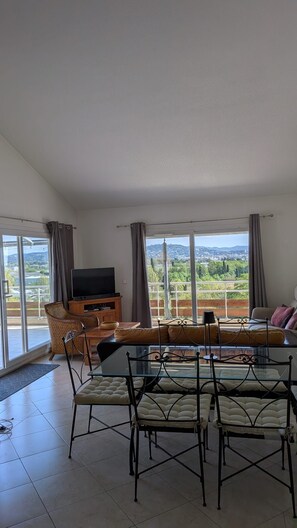 A spacious living room with a glass dining table, comfortable seating, a flat-screen TV, and a balcony with a scenic view.