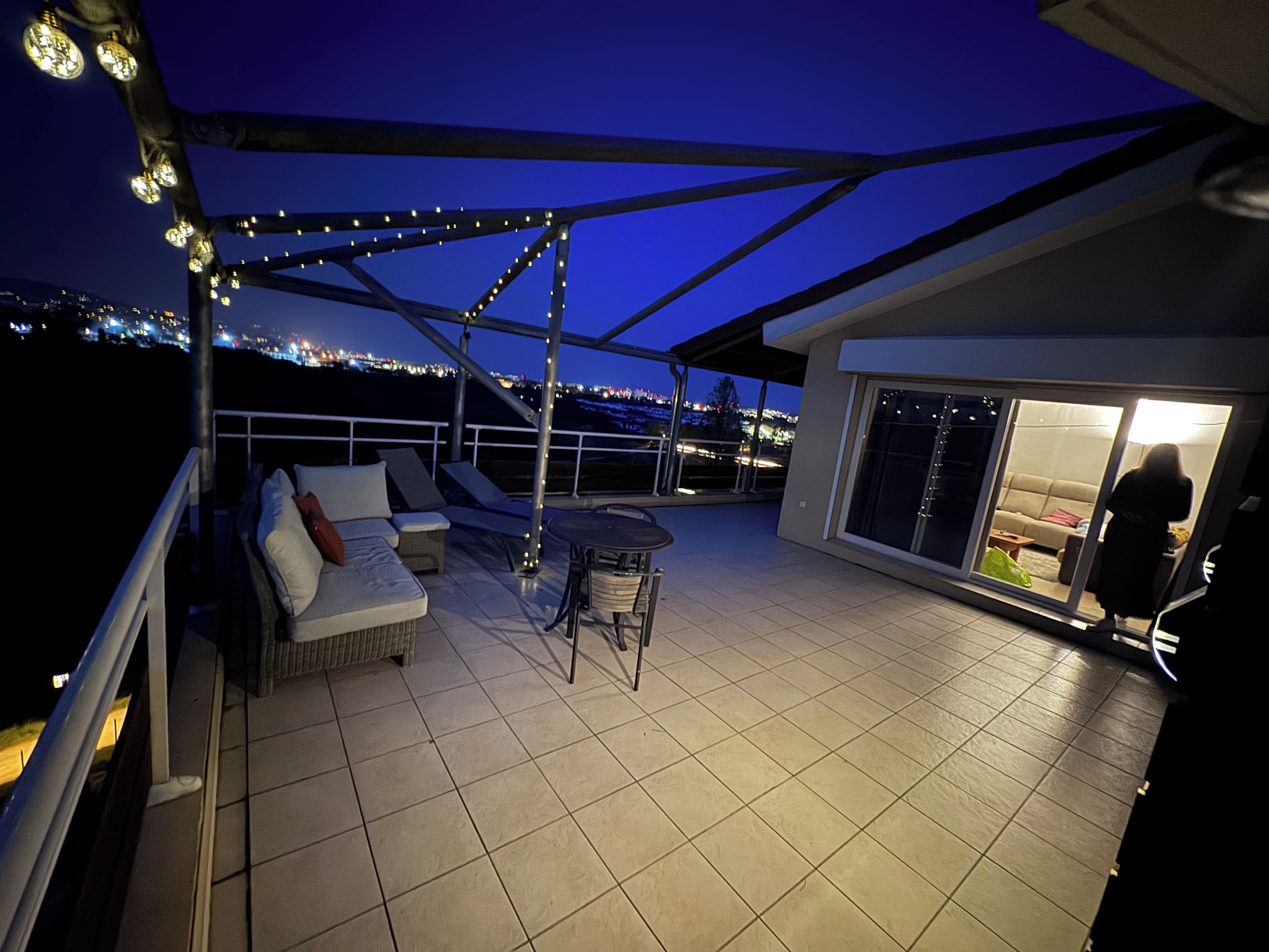 A rooftop terrace with a seating area, a table, and a view of the city lights at night.