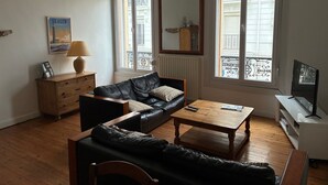 Un salon comprenant un canapé, une table basse, un téléviseur et une fenêtre donnant sur des immeubles.
