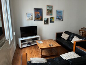 Uma sala de estar com televisão, mesa de centro de madeira, sofá de couro preto e vários cartazes emoldurados na parede.