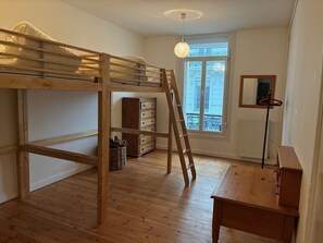 Quarto com cama de solteiro e beliche, escrivaninha de madeira, cômoda, cesta de vime, escada e janela com vista.