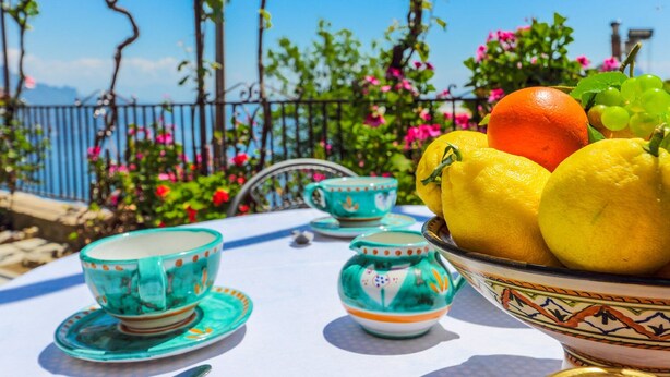 Una mesa dispuesta con fruteros, tazas de té y platillos, mostrando una vista de un jardín y una cerca.