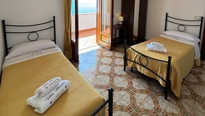 Habitación de hotel con dos camas, vista al mar a través de una puerta abierta, un ventilador de techo y una mesita con una lámpara.