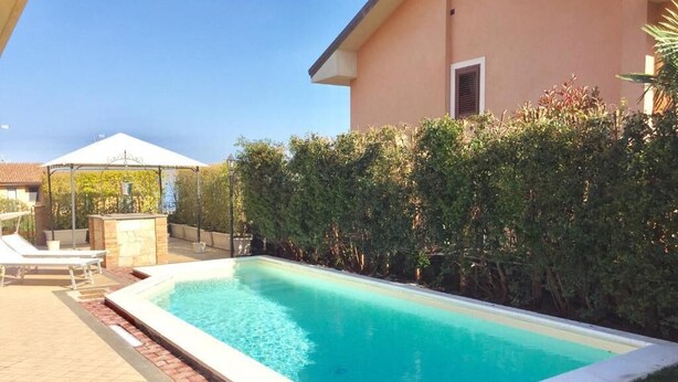 Ein rechteckiger Pool mit einer gefliesten Liegefläche, umgeben von einer niedrigen Hecke und einem Gebäude mit Satteldach.