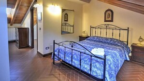 Ein Schlafzimmer mit schrägem Dach, einem Bett mit blumigem, blauem Überwurf, einem Nachttisch mit Lampe und einem an der Wand befestigten Spiegel.