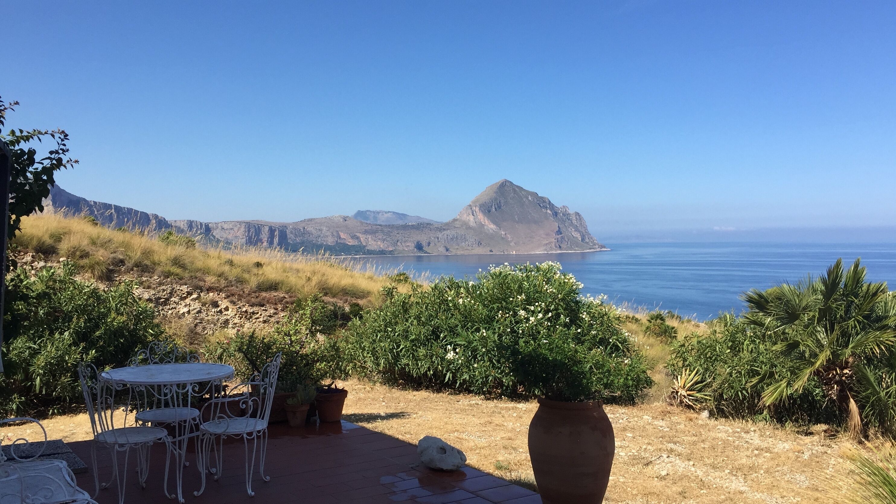 A patio with a table and chairs, surrounded by greenery and overlooking a coastal landscape with mountains and the sea.