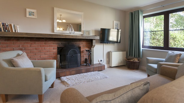 A living room with a fireplace, two sofas, a television, and a window with curtains. - Burnham A -  a self catering cottage that sleeps 3 guests  in 1 bedroom (Holt)