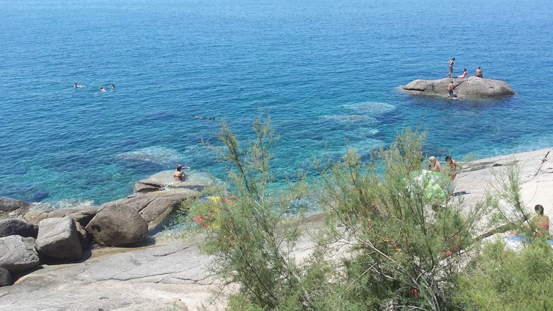 A coastal scene with clear blue water, rocky outcrops, and sparse vegetation.
