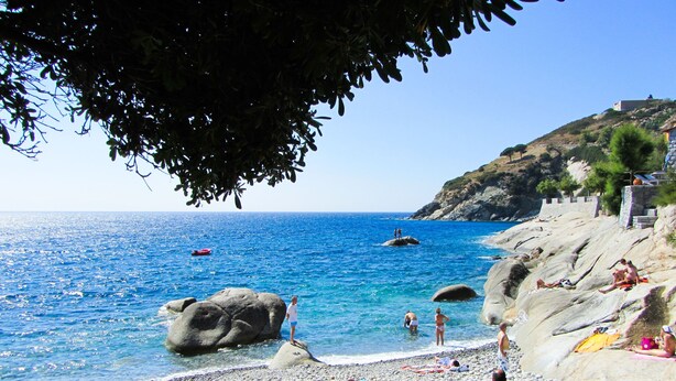 A pebble beach with clear blue water, rocky terrain, and a few people sunbathing. - Charming and cozy little house in the center of Pomonte, just steps from the sea (Pomonte)