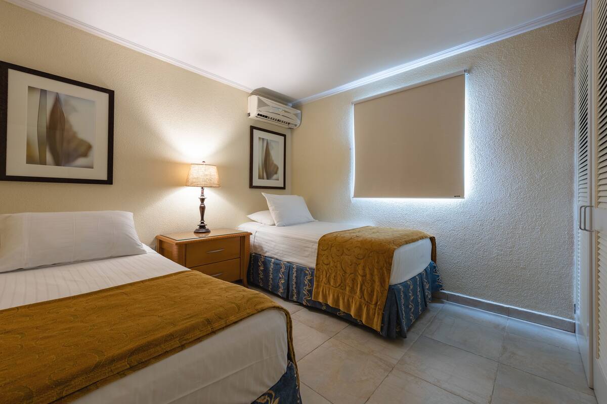 A hotel room with two beds, a nightstand, a lamp, an air conditioner, a window, and a framed picture on the wall.