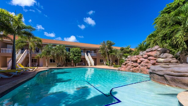 Une piscine à l’eau cristalline, entourée de palmiers et bordée d’un bâtiment dont le toit est recouvert de tuiles rouges.