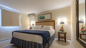 Une chambre d’hôtel comprenant un lit, deux tables de chevet, un climatiseur fixé au mur, un tableau encadré et un miroir agrémenté d’un arrangement floral.