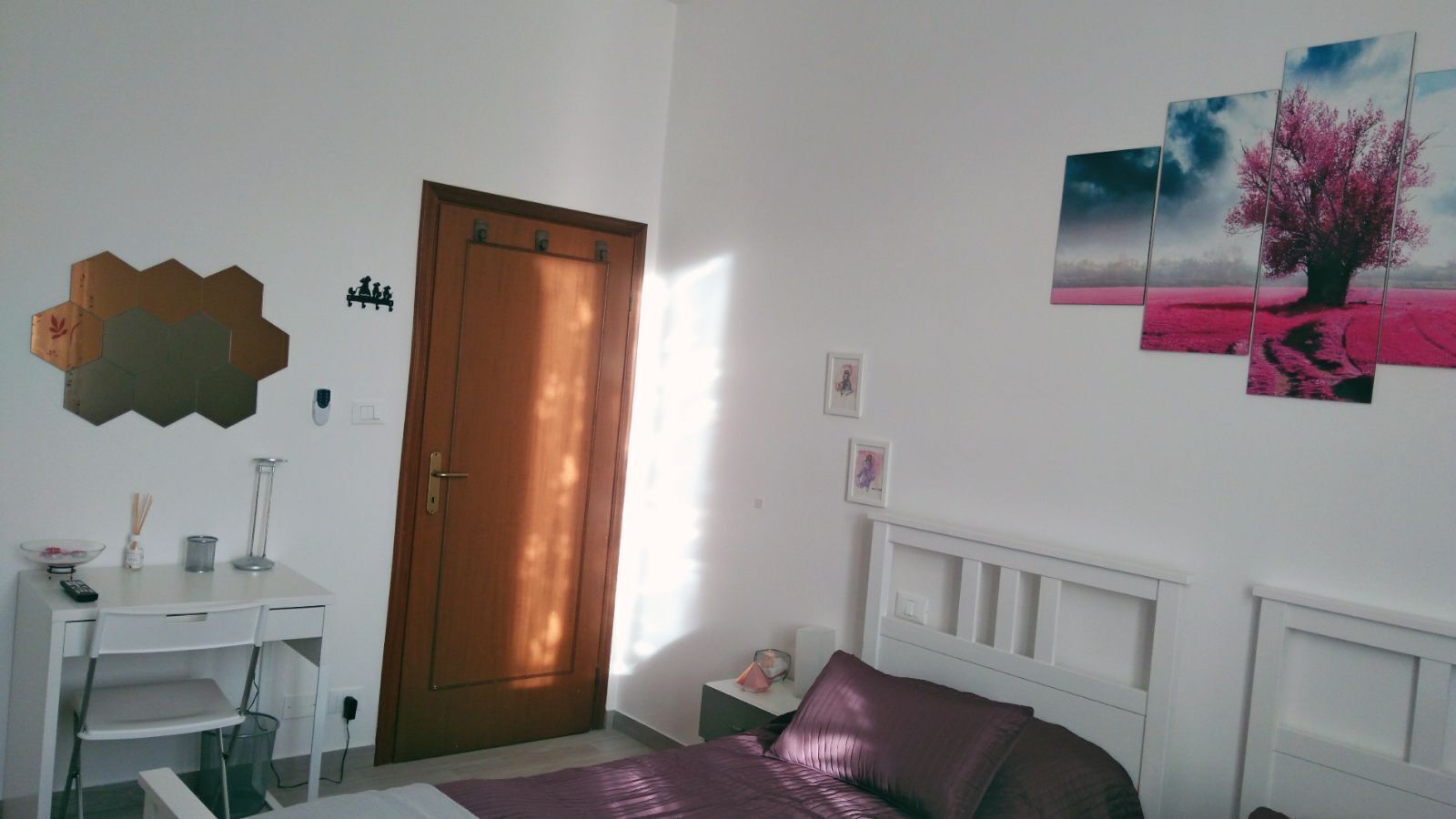Een slaapkamer met een houten deur, een wit nachtkastje, een bed met een paarse deken en een muur met een ingelijst beeld van een boom.