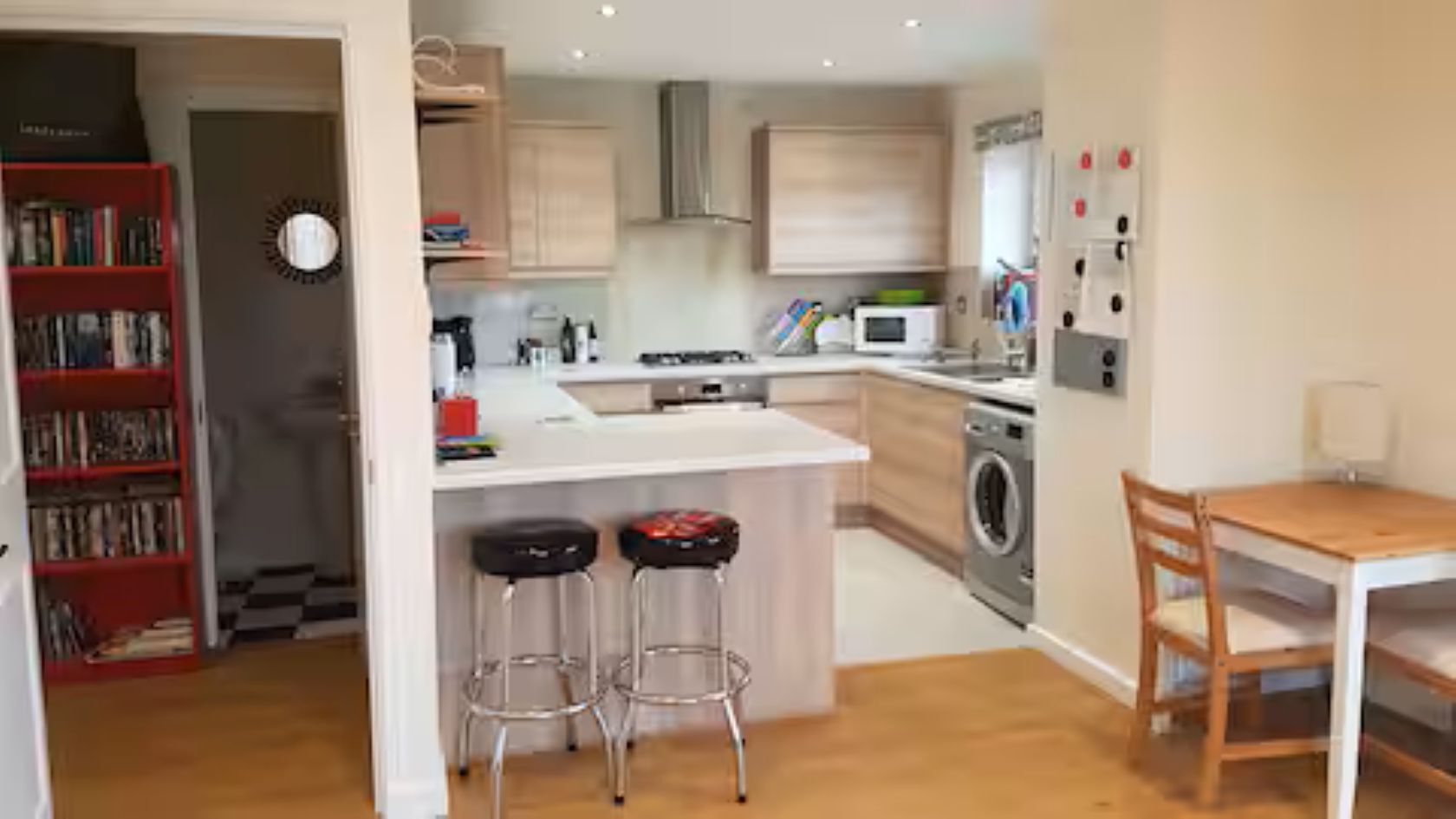 A compact kitchen with a central island, bar stools, a dining table, and a bookshelf.