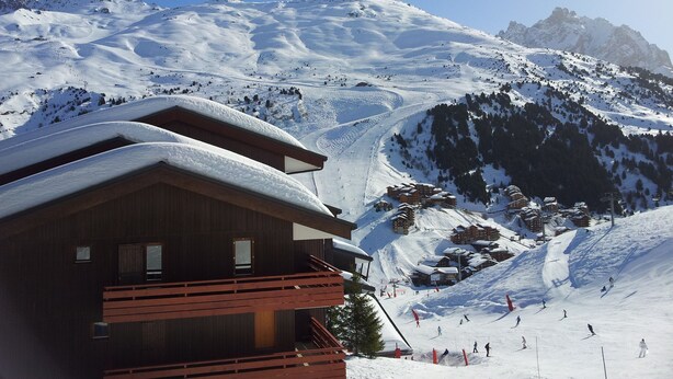 A wooden chalet with a balcony, set against a snowy mountain backdrop.