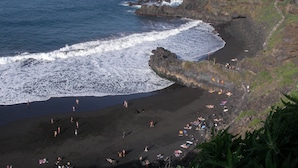 Eine Küstenlandschaft mit felsigem Ufer, einem Strand mit sonnenden Personen und einem Pfad, der den Klippen hinaufführt.