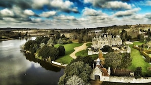 Un château historique doté d’une vaste cour, avec une rivière au premier plan et une végétation luxuriante tout autour.