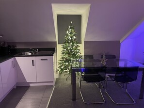 Eine moderne Küche mit weißen Schränken, einer schwarzen Arbeitsplatte und einem Glastisch mit zwei schwarzen Stühlen. Eine kleine, beleuchtete Weihnachtstanne steht in der Ecke.