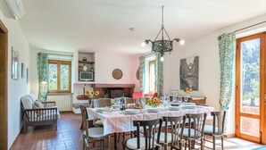 Un comedor con una mesa redonda dispuesta para seis personas, rodeada de sillas de madera. Se ve una chimenea al fondo y ventanas con cortinas.