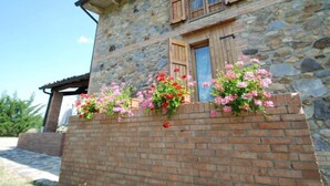 Un edificio de piedra con contraventanas de madera, una base de ladrillo y jardineras con flores rosadas y rojas.
