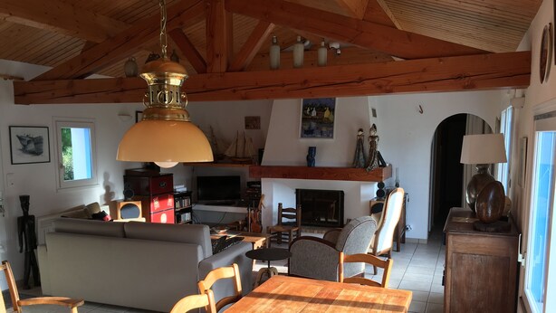 Amplia sala de estar con una mesa de comedor de madera, sillas, un sofá y una chimenea. Se observa una zona de cocina con un televisor y varios objetos en los estantes.