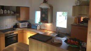 Una cocina moderna con armarios de madera, horno empotrado, microondas y un lavamanos. Hay una ventana que ofrece luz natural y vistas a la vegetación exterior.