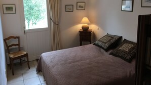 Un dormitorio con cama, una silla, una mesita de noche con lámpara, ventana con cortinas y cuadros en la pared.