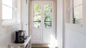 Ein helles, weißes Interieur mit einer Doppeltür, die zu einem Außenbereich führt, einer Kaffeemaschine auf einem Regal und einem kleinen Blumentopf auf einem Fensterbrett.