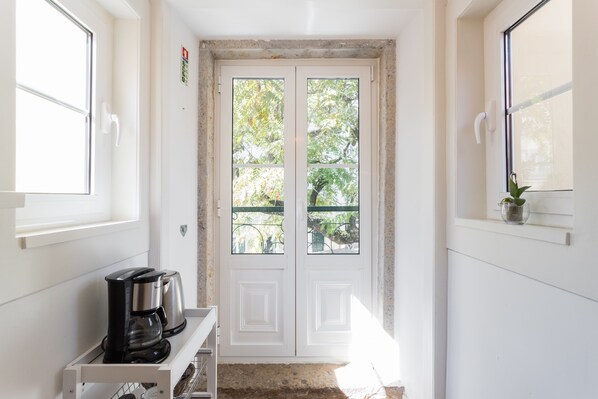 A clean, white interior with a double door leading to an outdoor area, a coffee maker on a shelf, and a small potted plant on a window sill.