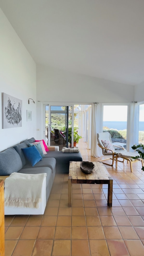 Uma sala de estar espaçosa com um sofá cinza grande, uma mesa de centro e vista para o oceano através de amplas janelas.