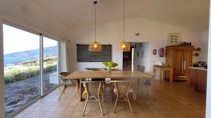 Uma sala de jantar com mesa de madeira e cadeiras de vime, vista para o mar através de amplas janelas, e uma cozinha com armários de madeira.