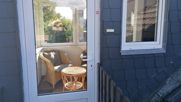 Une porte avec un panneau de verre, une chaise en osier, une petite table ronde et une fenêtre ornée d’une plaque nominative.