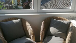 Une pièce avec deux fauteuils en osier et une petite table ronde, une fenêtre donnant sur un jardin et des bâtiments, et un mur blanc avec une prise de courant.