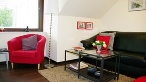 Un salon avec un fauteuil rouge, un canapé en cuir noir, une table basse en verre et un lampadaire. Deux tableaux encadrés ornent le mur, et une fenêtre offre une vue sur la verdure.