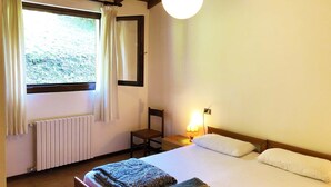 Ein Schlafzimmer mit Bett, Sessel, Nachttisch mit Lampe, Fenster mit Vorhängen und Heizkörper.