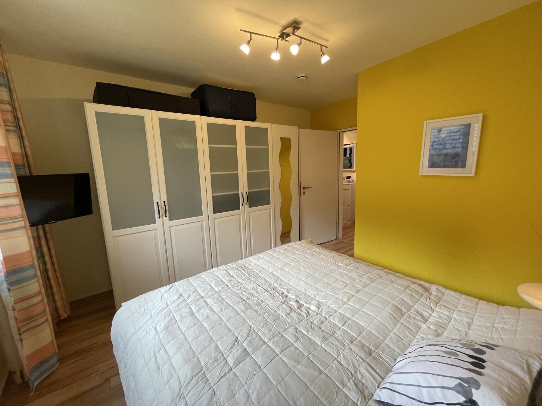 Una camera da letto con un letto, un armadio con ante in vetro, una televisione a parete e un quadro appeso.