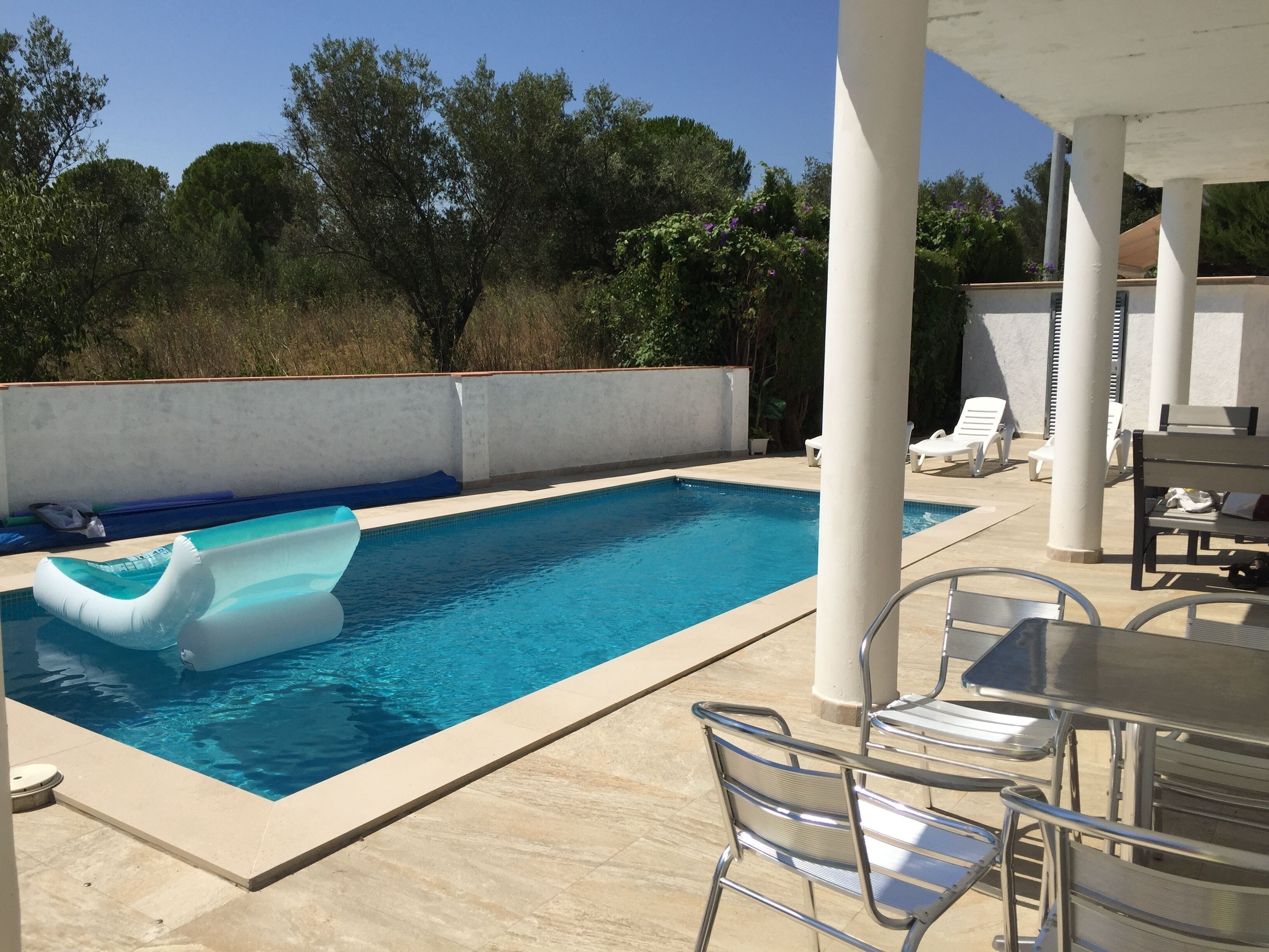 Una piscina con agua azul cristalina, rodeada de columnas blancas y sillones de descanso.