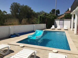 A rectangular swimming pool with a blue inflatable slide, surrounded by white lounge chairs and a covered area with a roof. - Spacious holiday home with private pool, internet, air conditioning (Roses)