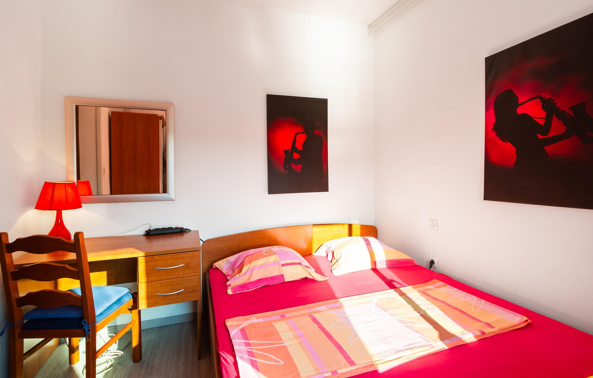 A bedroom with a bed, a desk with a chair, a lamp, and two wall art pieces.