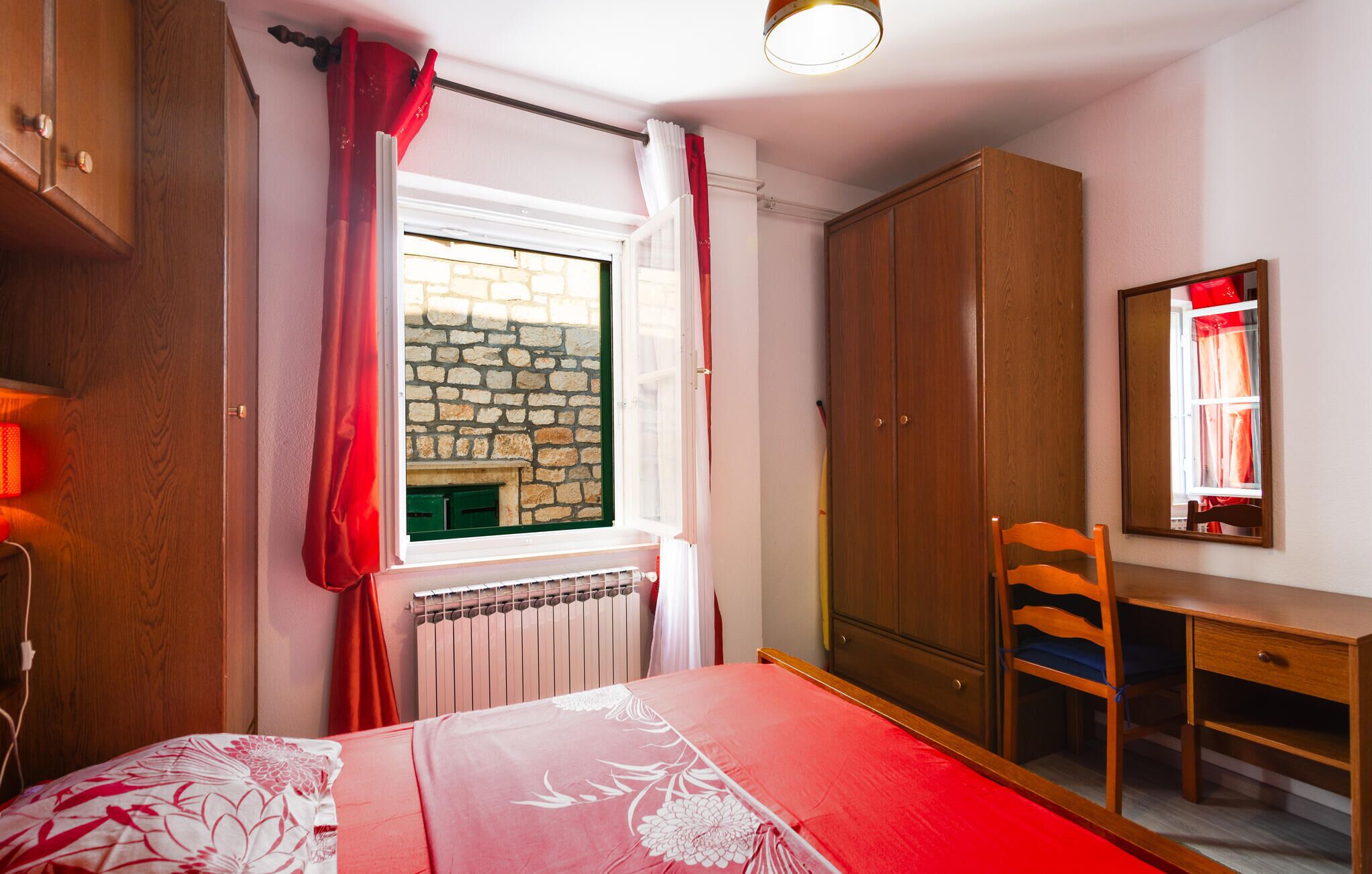 A bedroom with a bed, a wooden wardrobe, a desk, a chair, a window with curtains, and a radiator.