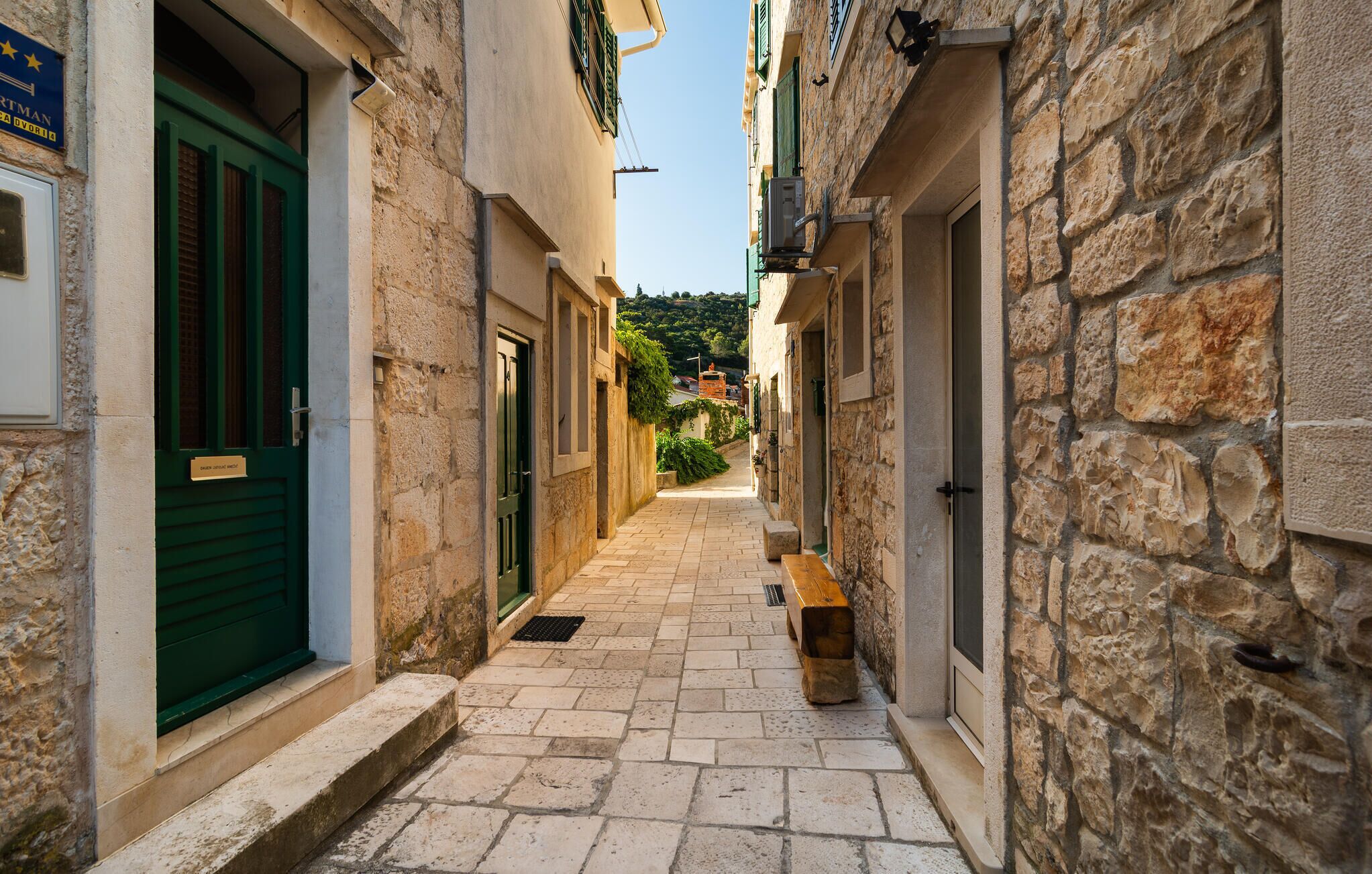 A narrow stone-paved alleyway between buildings with green shutters and a bench.