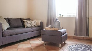 Ein Wohnzimmer mit grauem Sofa, einem kleinen Hocker und einer dekorativen Vase auf einem Fensterbrett.
