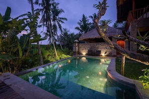 A swimming pool with a stone wall and thatched roof structure, surrounded by tropical vegetation. - ILA Kitchen & Villa (Ubud)