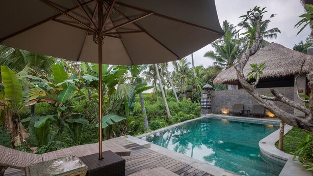 Área à beira da piscina com espreguiçadeiras e um guarda-sol, cercada por vegetação tropical exuberante e uma estrutura com telhado de palha.